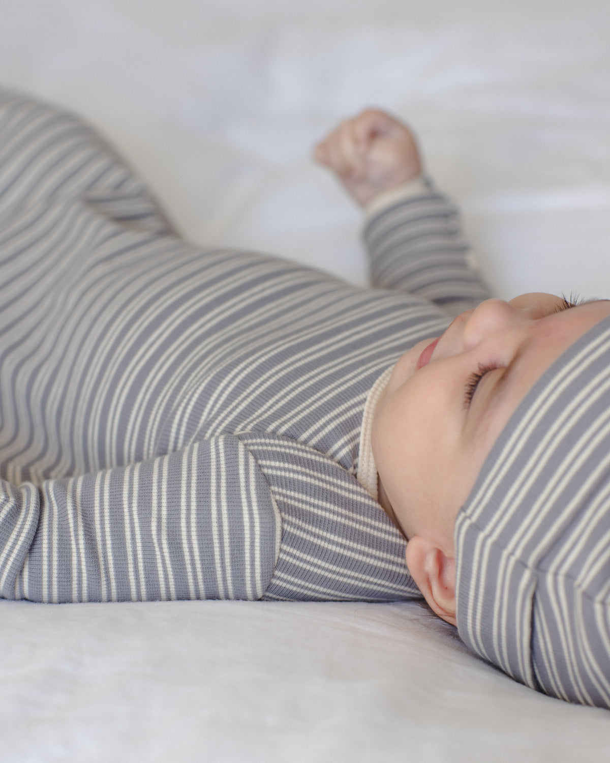 A baby lying on a white blanket, wearing a gray and white striped long-sleeve footless sleeper and a matching striped bonnet.