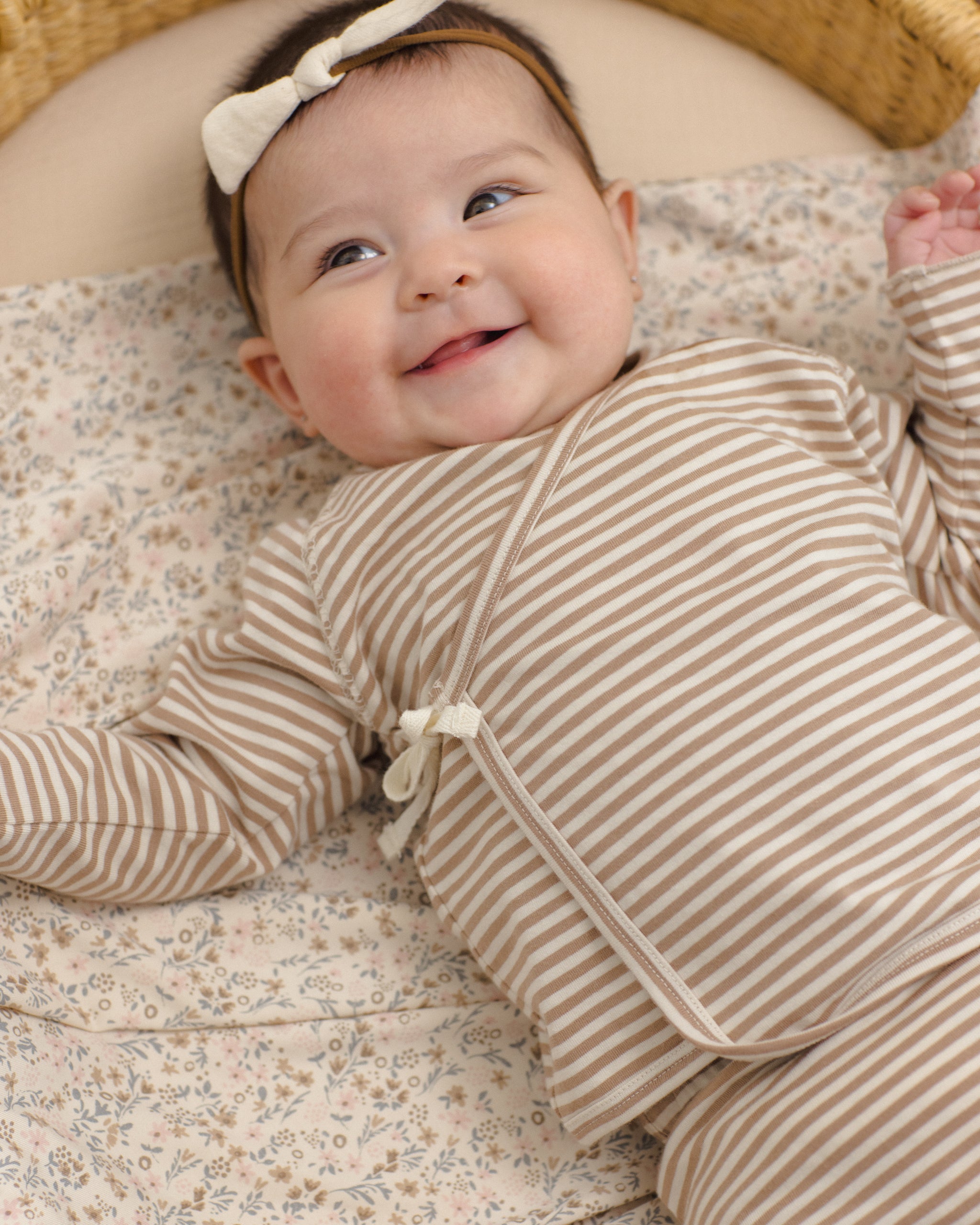 A smiling baby lying on a floral blanket, wearing a beige and white striped long-sleeve romper with a tie closure and a matching cream bow headband.