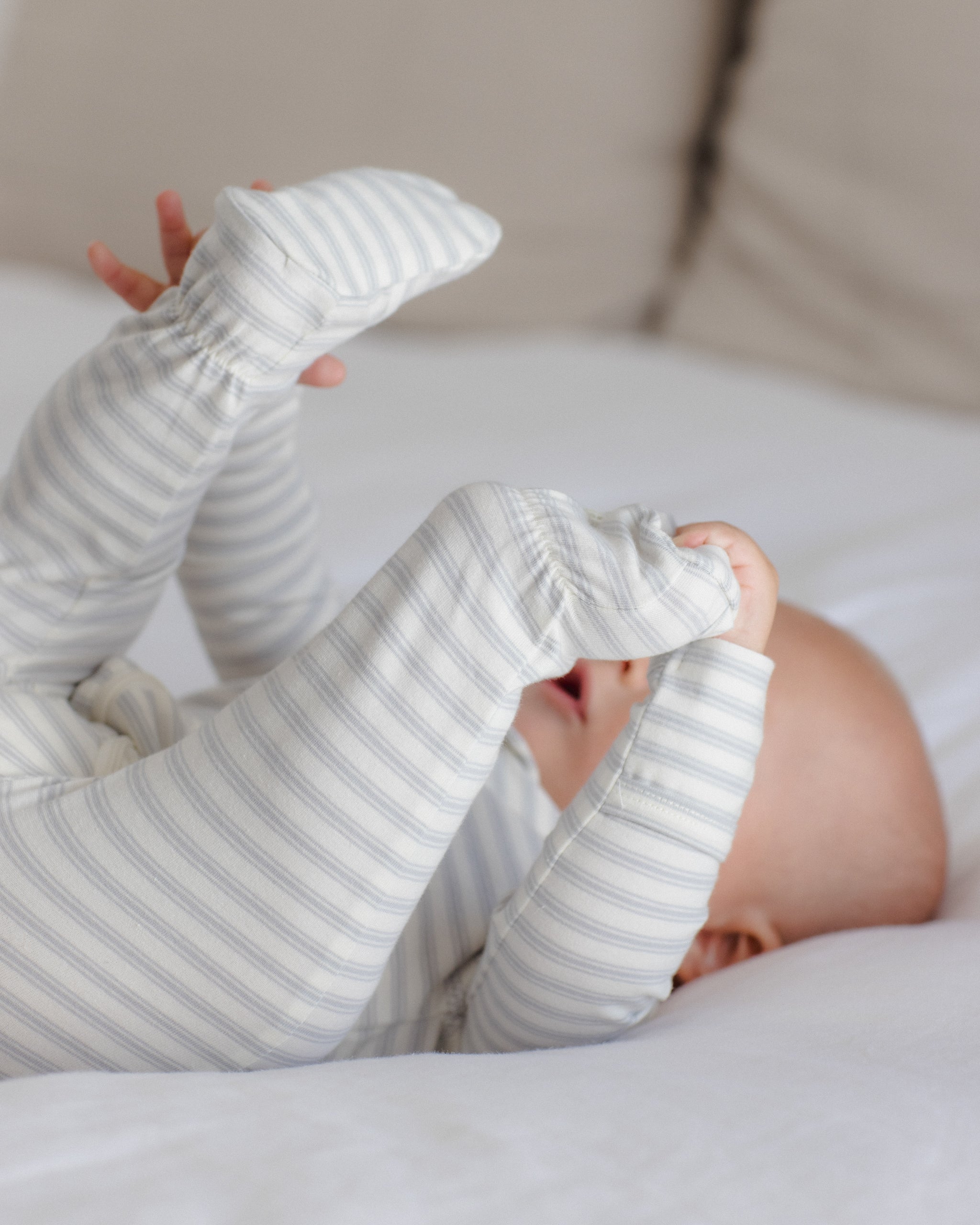A baby lying on a white bed, wearing a long-sleeve footed sleeper with thin gray and white stripes.