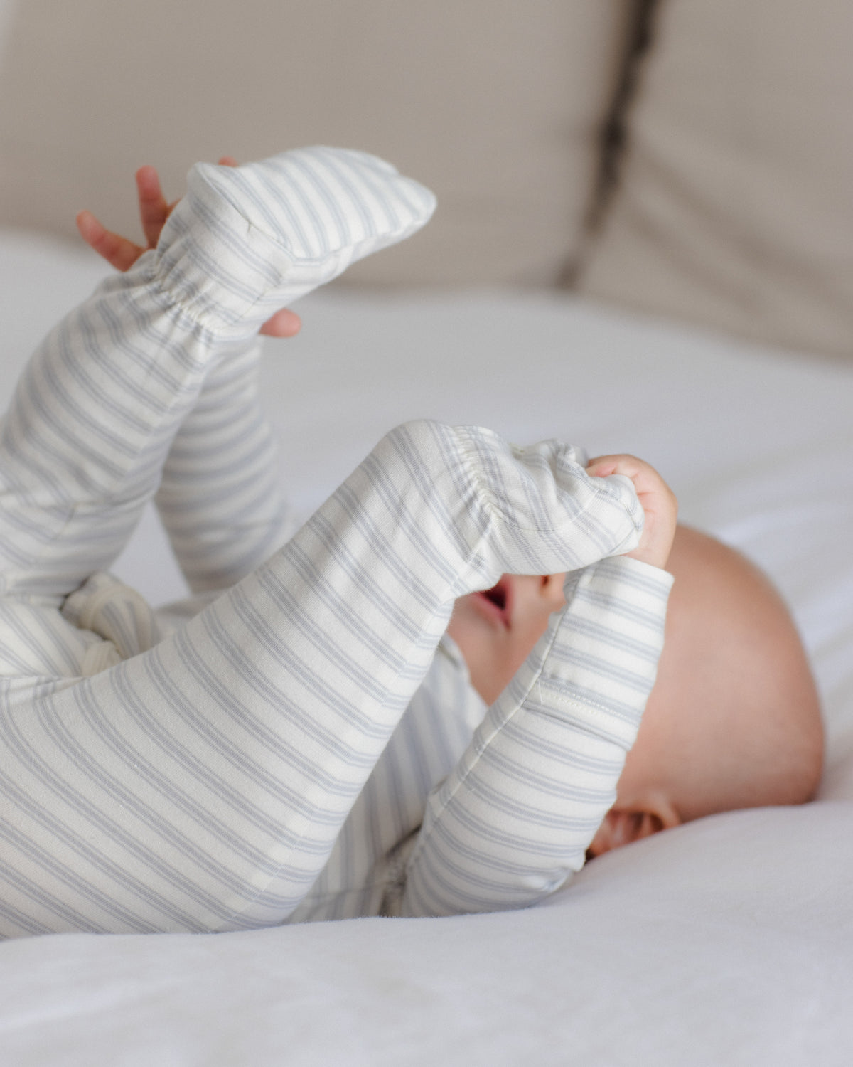 A baby lying on a white bed, wearing a long-sleeve footed sleeper with thin gray and white stripes.
