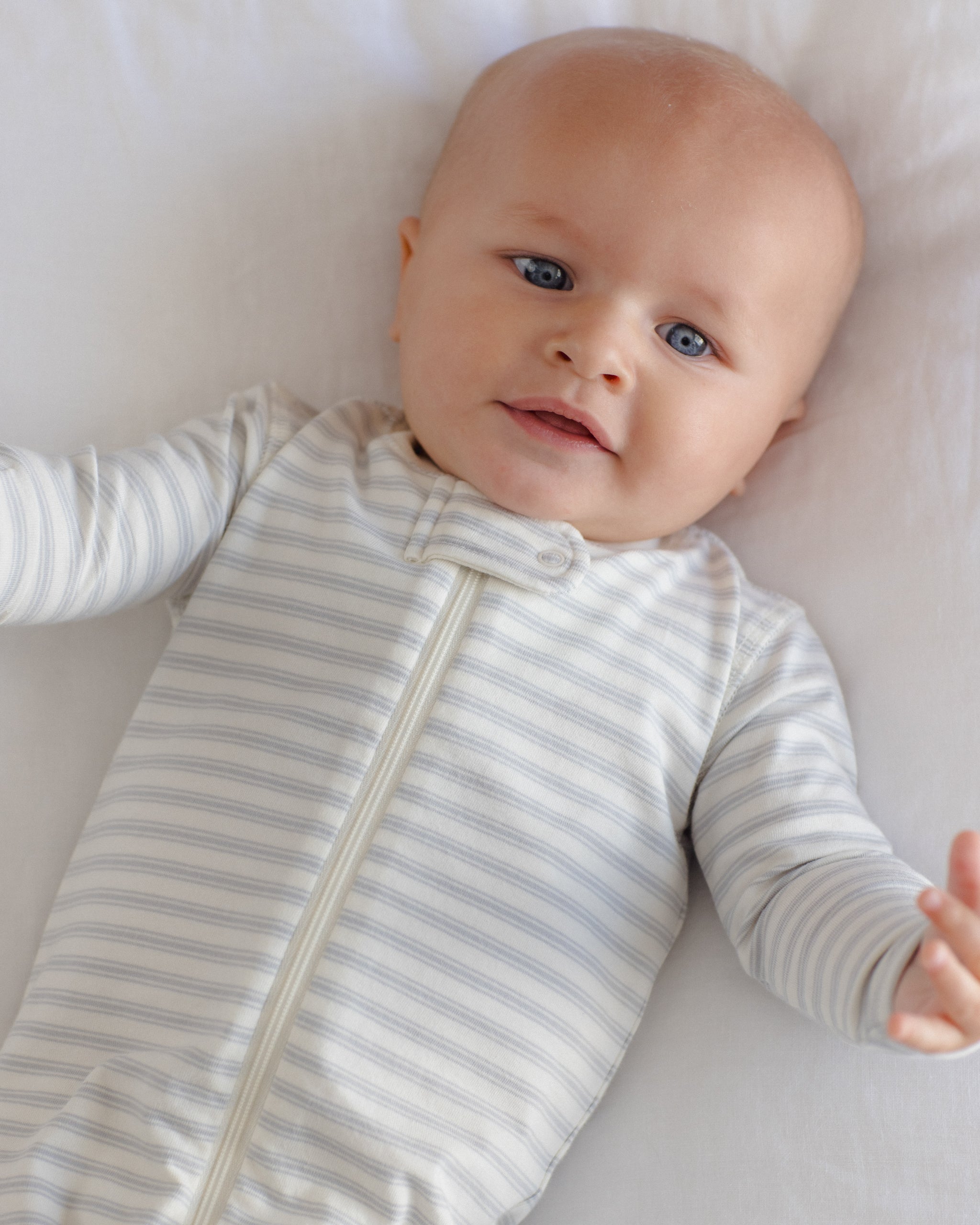 A baby lying on a white blanket wearing a long-sleeve zip-up sleeper with thin gray and white stripes.