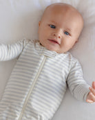 A baby lying on a white blanket wearing a long-sleeve zip-up sleeper with thin gray and white stripes.