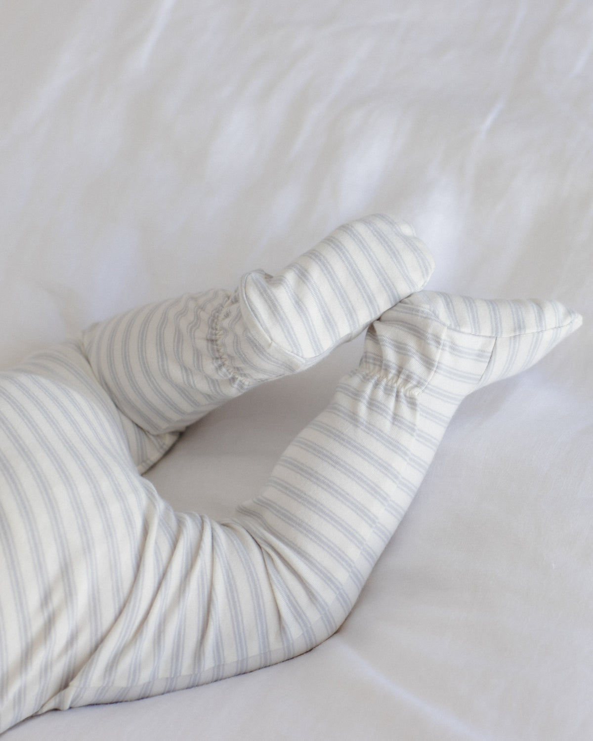 A close-up of a baby's legs wearing soft, cream-colored footed socks or booties with thin gray stripes, resting on white bedding.