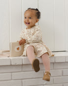 A smiling toddler girl sits on a white brick ledge holding a small beige book. She is wearing a cream long-sleeve dress with a delicate floral pattern, pink tights, and tan Mary Jane shoes against a white paneled wall background.