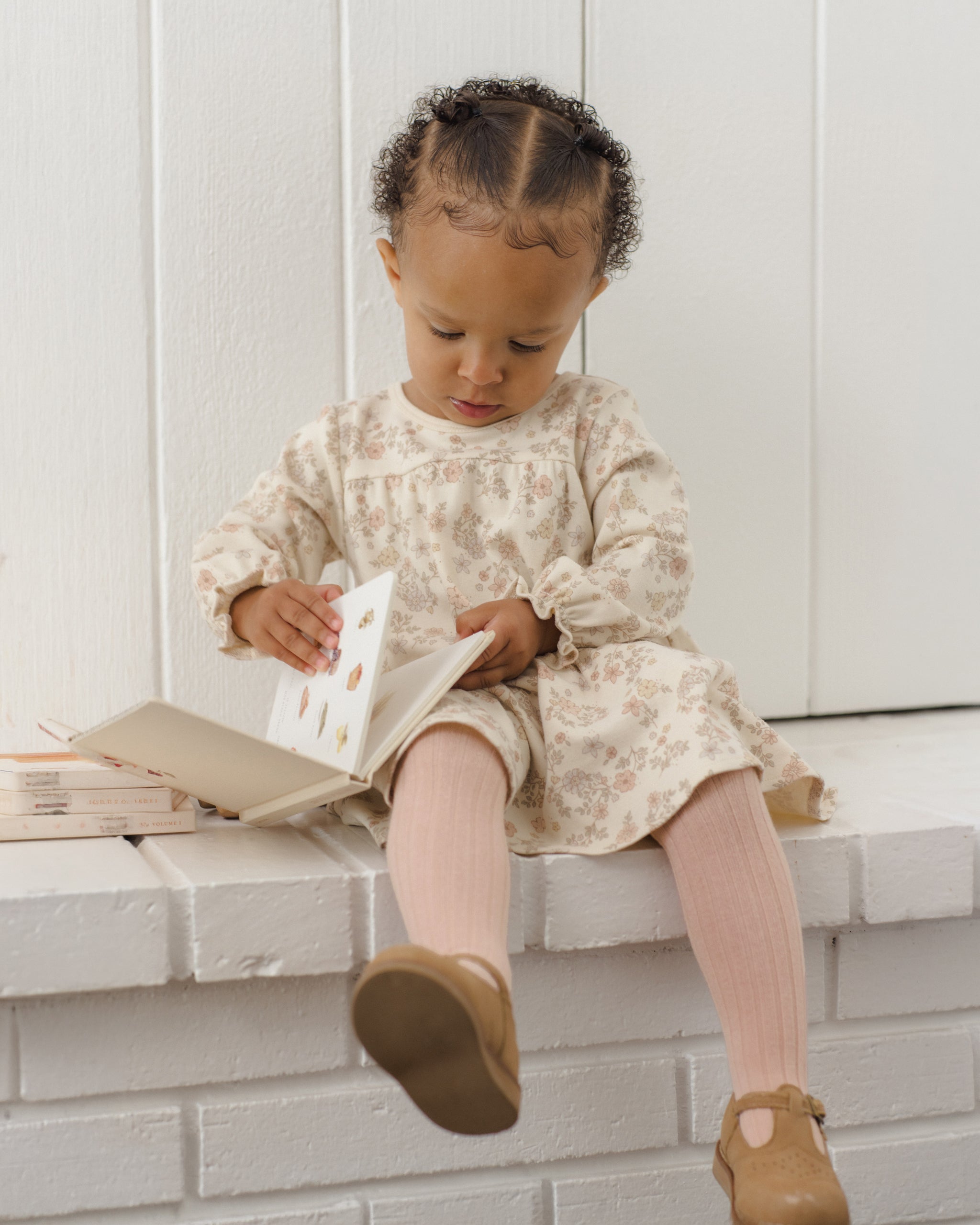 A toddler girl sits on a white brick bench reading a book, wearing a cream floral long-sleeve dress, pink ribbed tights, and brown Mary Jane shoes.