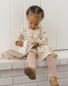 A toddler girl sits on a white brick bench reading a book, wearing a cream floral long-sleeve dress, pink ribbed tights, and brown Mary Jane shoes.
