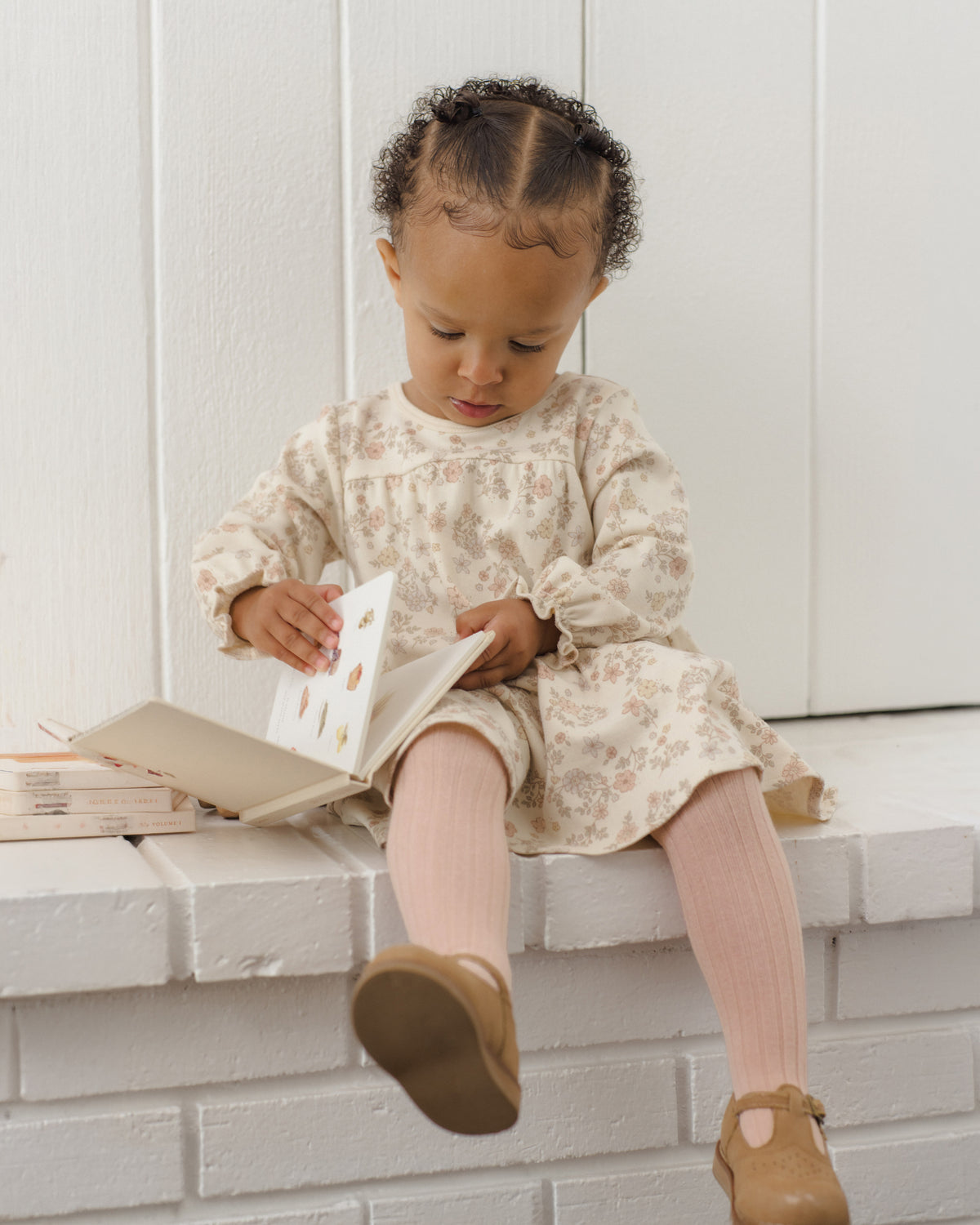 A toddler girl sits on a white brick bench reading a book, wearing a cream floral long-sleeve dress, pink ribbed tights, and brown Mary Jane shoes.