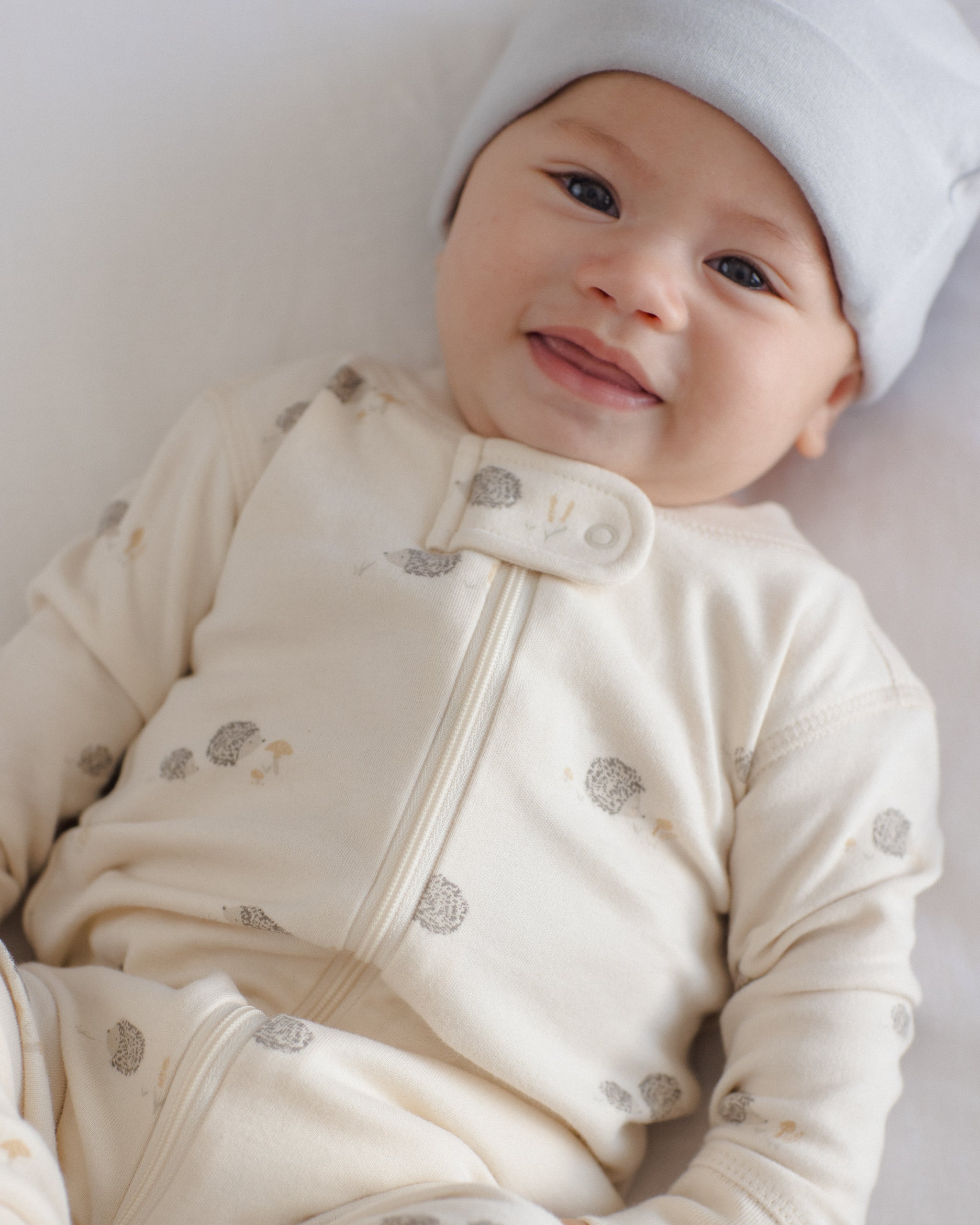 A smiling baby lying down wearing a cream-colored long-sleeve zip-up sleeper with small gray and beige mushroom prints, along with a light blue beanie.