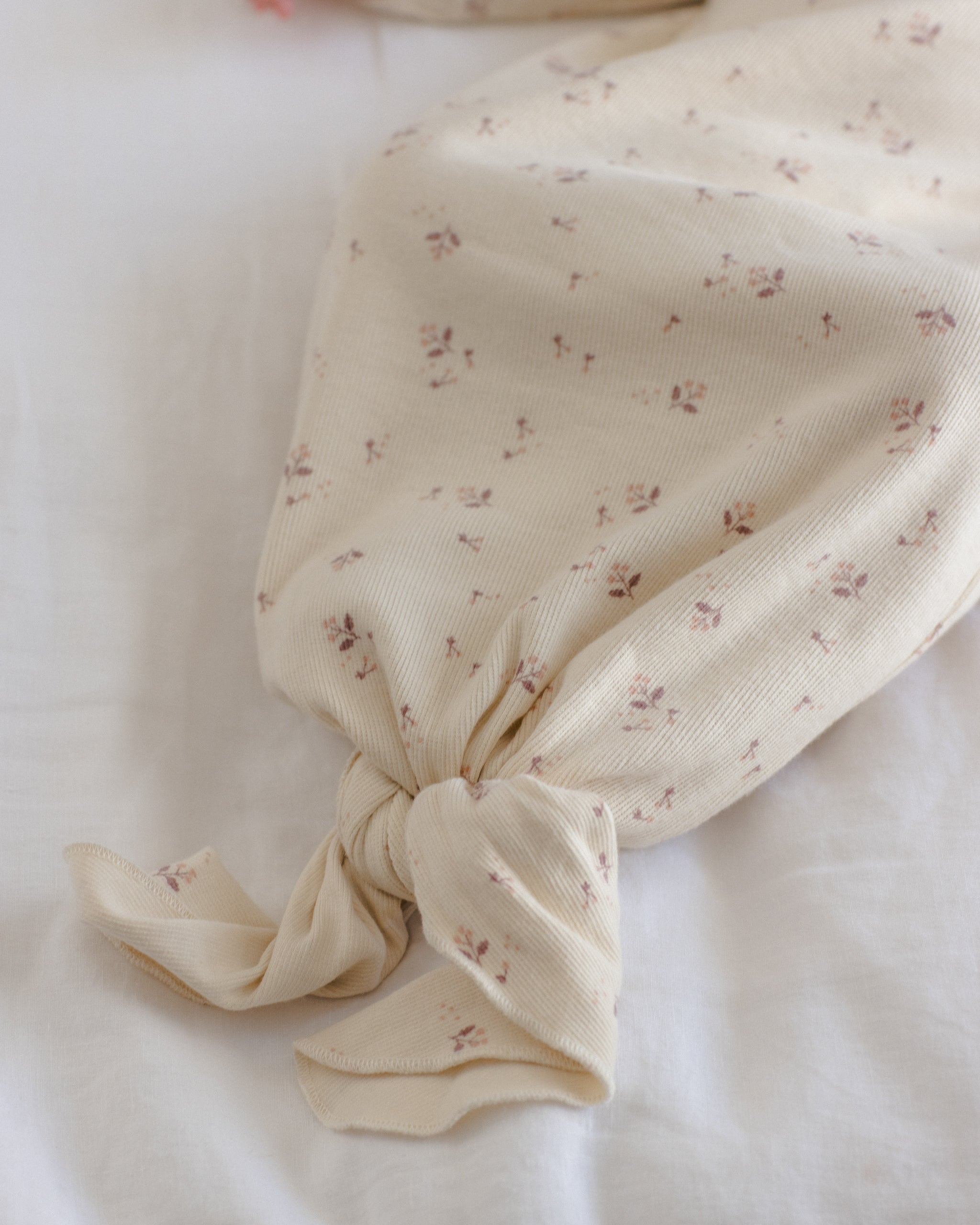 A cream-colored muslin fabric item, possibly a baby blanket or swaddle, featuring a delicate all-over floral print in muted pink and brown, tied neatly at the bottom on a soft white fabric background.