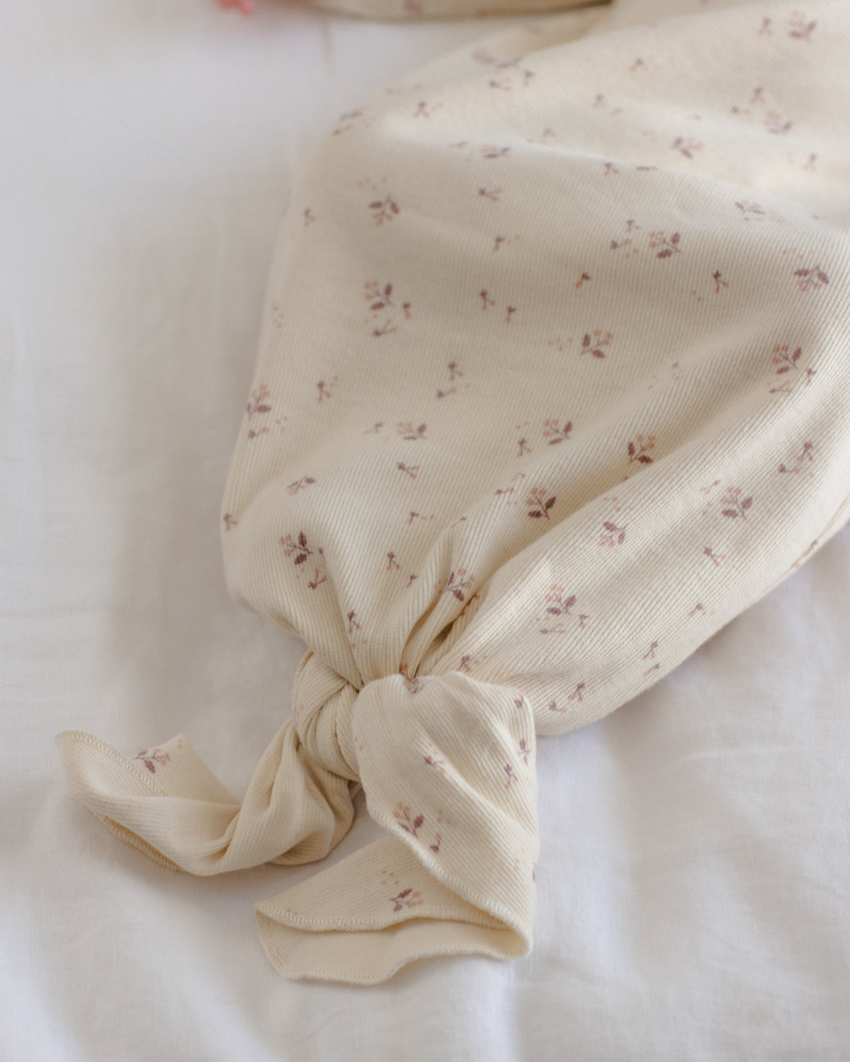 A cream-colored muslin fabric item, possibly a baby blanket or swaddle, featuring a delicate all-over floral print in muted pink and brown, tied neatly at the bottom on a soft white fabric background.