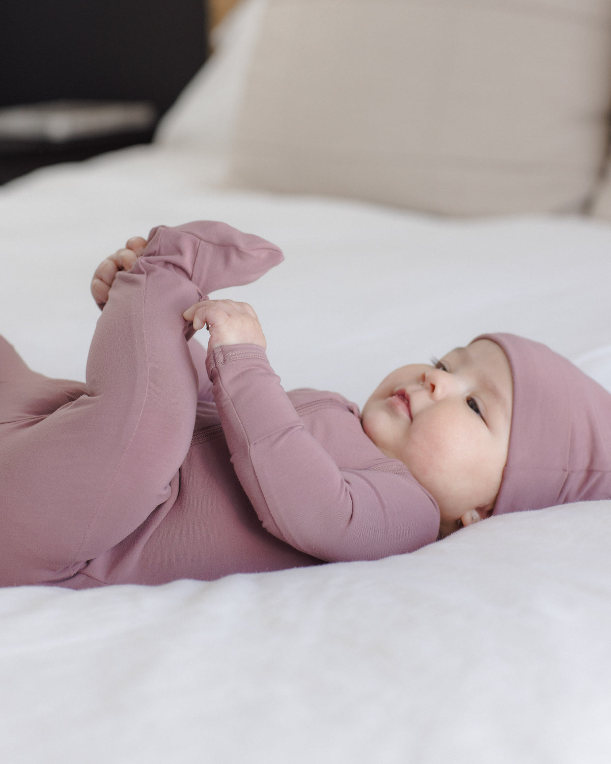 A baby lying on white bedding wearing a dusty mauve long-sleeve footed one-piece and matching hat.