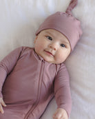 A baby lying on a white blanket, wearing a dusty rose pink long-sleeve zip-up sleeper and matching pointed knit hat.