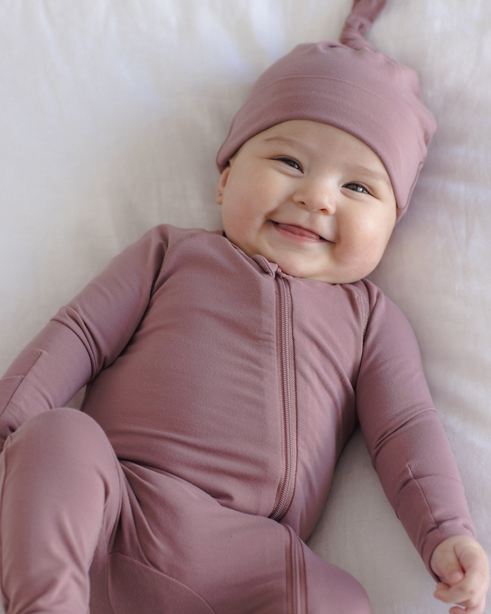 A smiling baby lying on a white blanket, wearing a dusty mauve zip-front footed onesie and matching pointed hat.