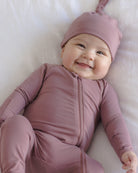 A smiling baby lying on a white blanket, wearing a dusty mauve zip-front footed onesie and matching pointed hat.