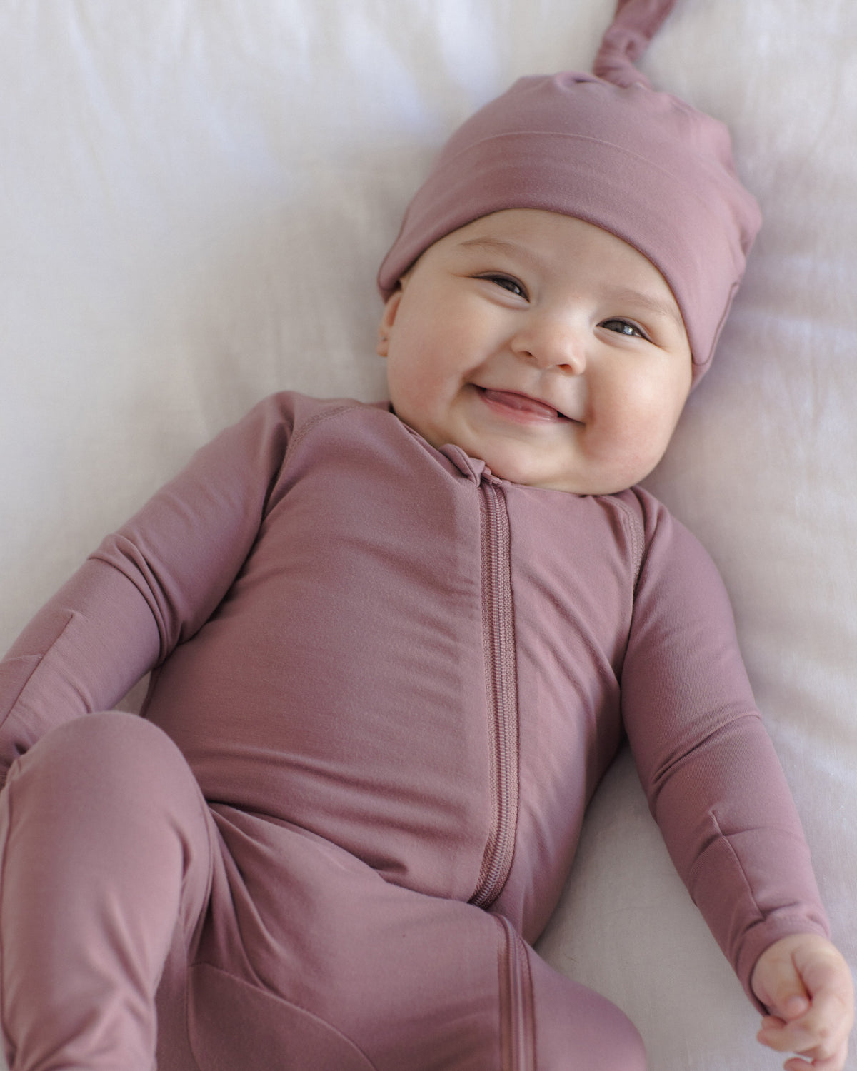 A smiling baby lying on a white blanket, wearing a dusty mauve zip-front footed onesie and matching pointed hat.