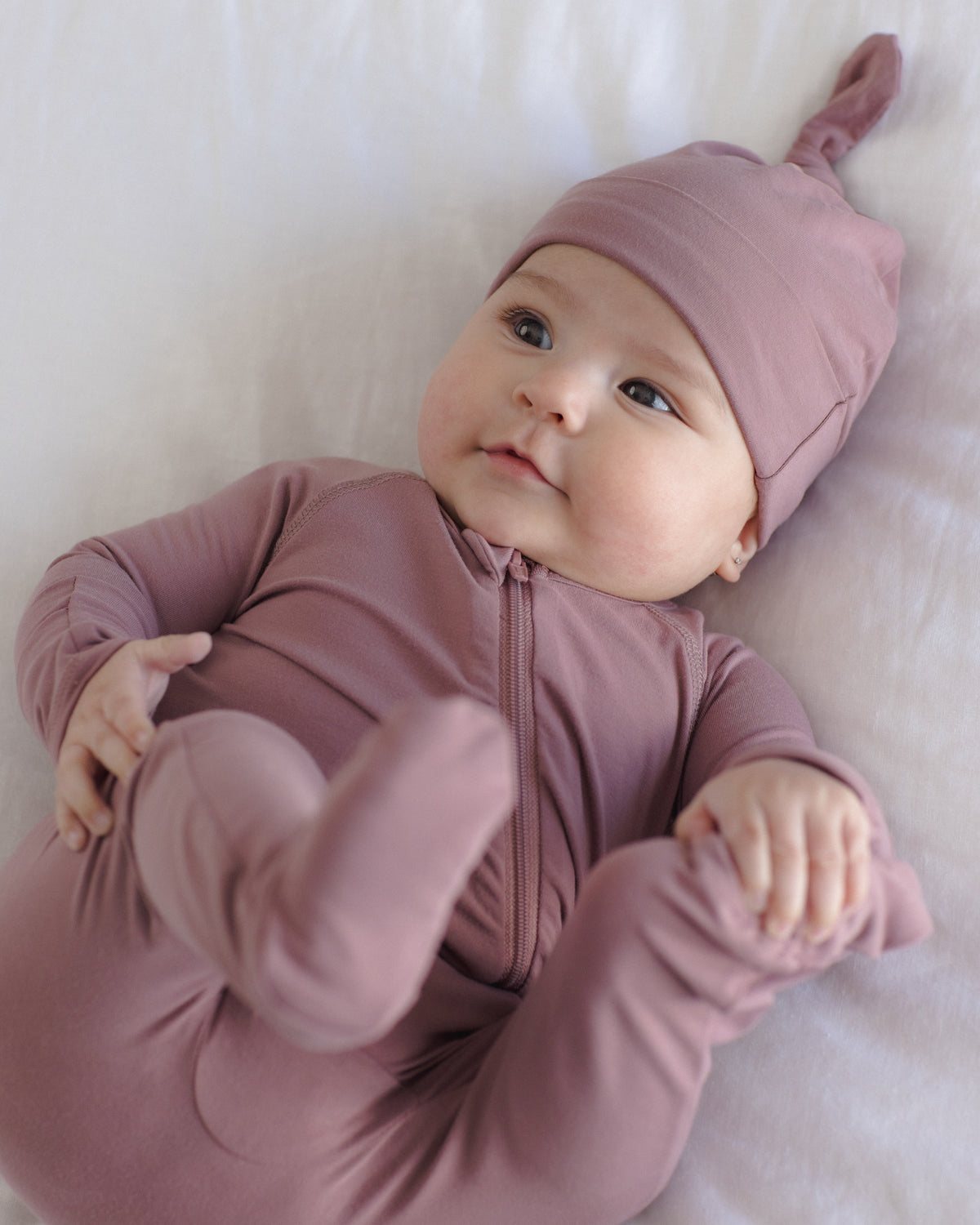 A baby lying on a white blanket, wearing a dusty mauve zip-up one-piece sleeper with a matching pointed knit hat.