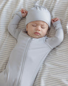 A sleeping newborn baby wearing a light gray long-sleeve zip-up sleeper and matching knotted beanie, lying on a beige and white striped blanket.