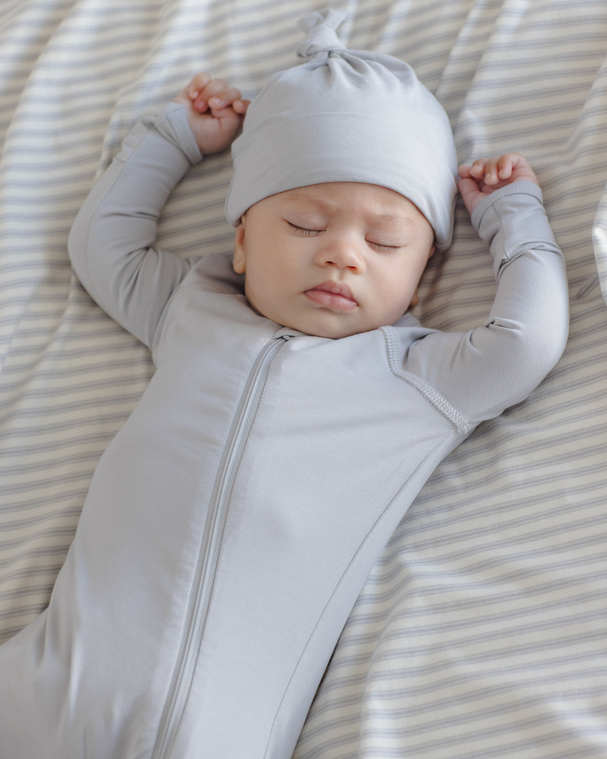 A sleeping newborn baby wearing a light gray long-sleeve zip-up sleeper and matching knotted beanie, lying on a beige and white striped blanket.