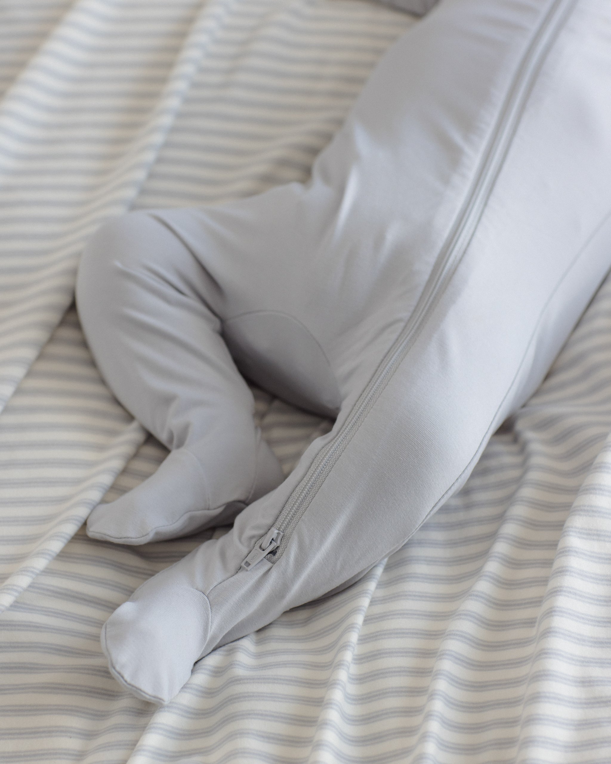 A close-up of a baby's legs and feet wearing a light gray footed sleeper with a zipper, lying on a cream and gray striped sheet.