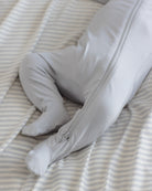 A close-up of a baby's legs and feet wearing a light gray footed sleeper with a zipper, lying on a cream and gray striped sheet.