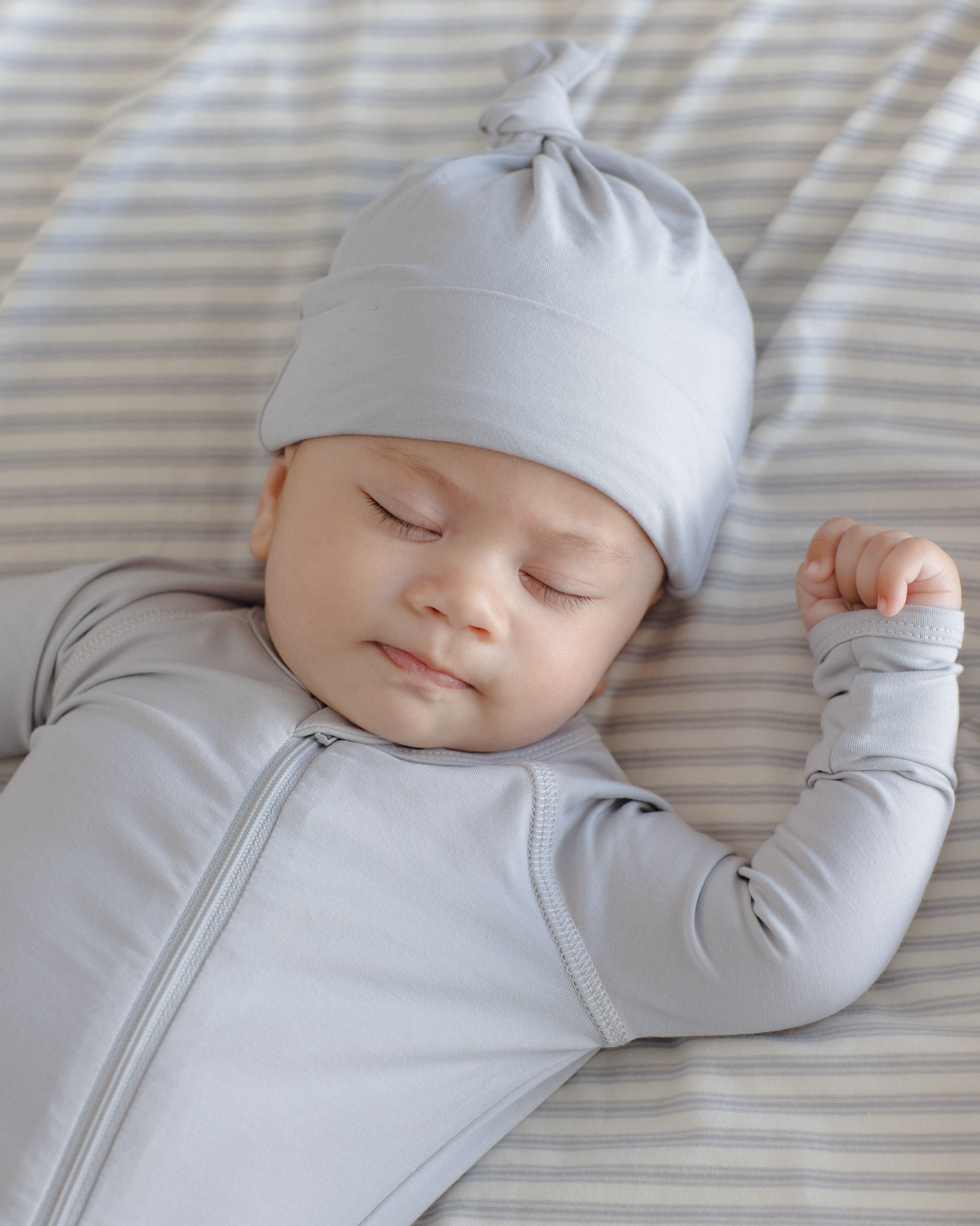 A sleeping baby wearing a light gray long-sleeve zip-up onesie and matching knotted beanie hat, lying on a striped bedding surface.