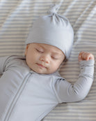 A sleeping baby wearing a light gray long-sleeve zip-up onesie and matching knotted beanie hat, lying on a striped bedding surface.