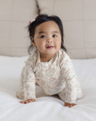 A baby girl wearing a cream-colored floral long-sleeve footless sleeper sits on a white bed, looking towards the camera.