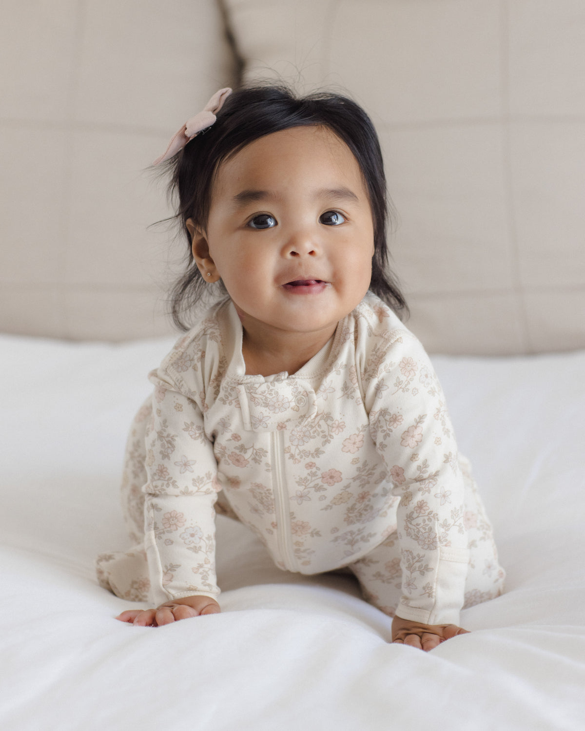 A baby girl wearing a cream-colored floral long-sleeve footless sleeper sits on a white bed, looking towards the camera.