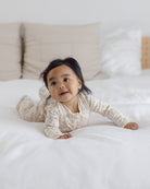 A baby wearing a cream and beige patterned long-sleeve romper lies on their stomach on a white bed with neutral pillows in the background.