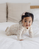 A baby crawling on a white bed wearing a cream-colored long-sleeve onesie with a delicate pink floral pattern and a matching pink bow in their hair.