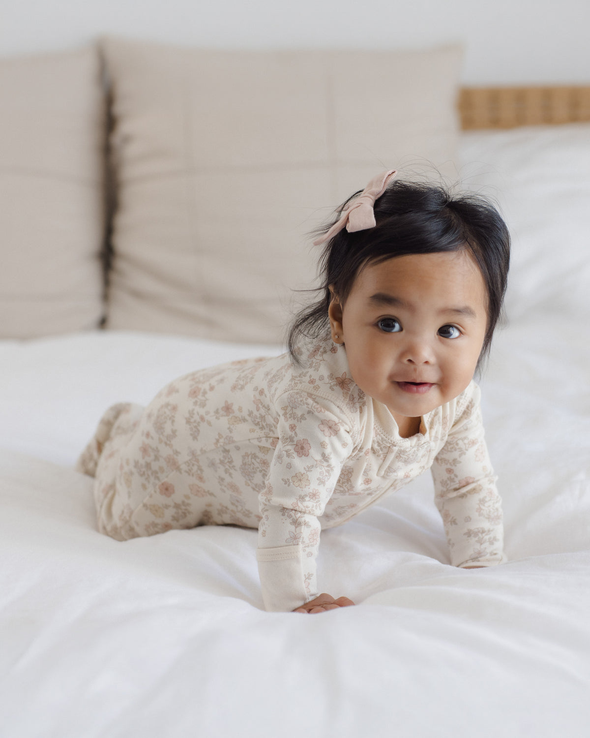 A baby crawling on a white bed wearing a cream-colored long-sleeve onesie with a delicate pink floral pattern and a matching pink bow in their hair.