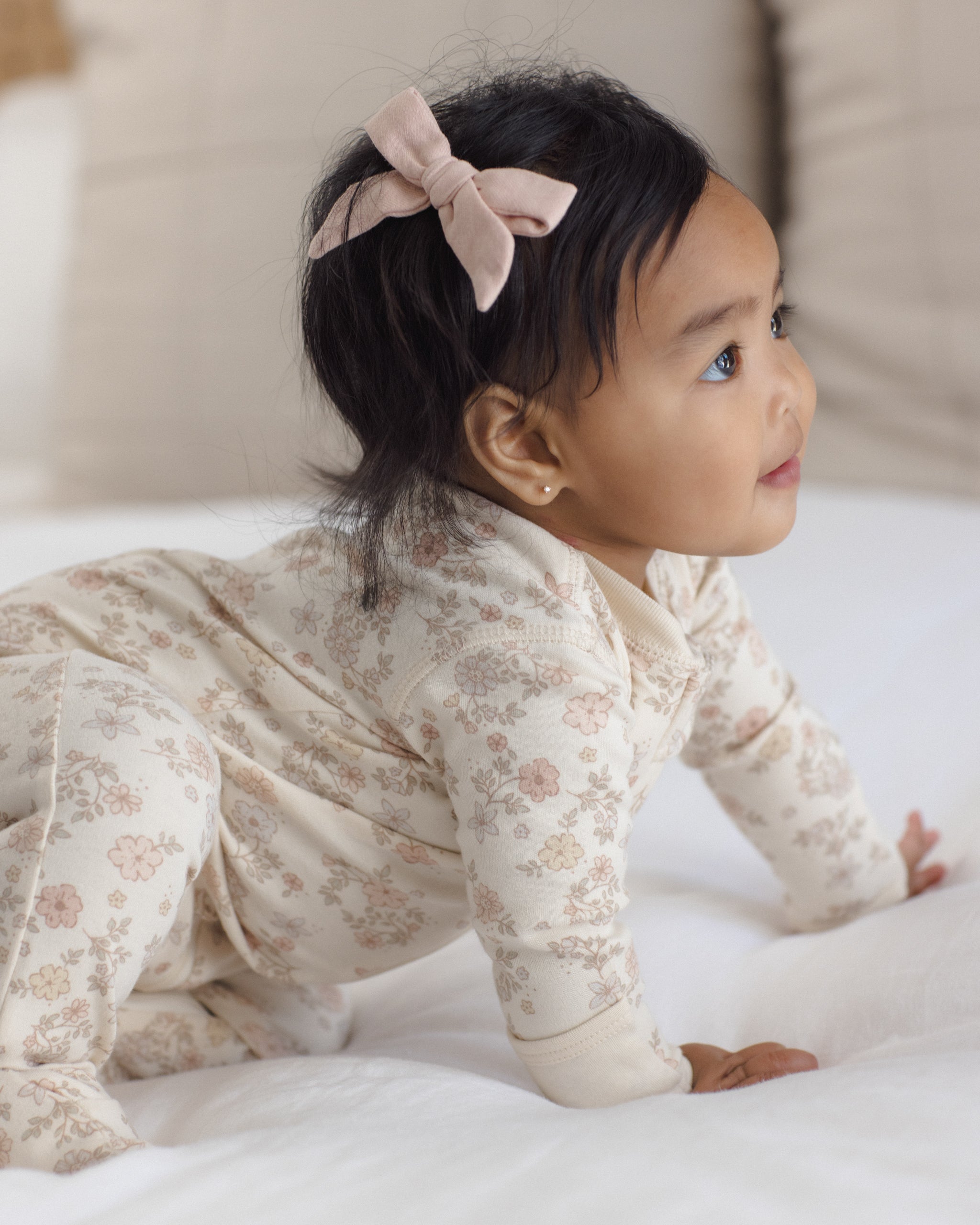 A baby crawling on a white bed wearing a cream-colored long-sleeve footed sleeper with a delicate pink floral pattern and a matching pink bow headband.