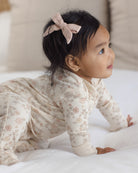 A baby crawling on a white bed wearing a cream-colored long-sleeve footed sleeper with a delicate pink floral pattern and a matching pink bow headband.