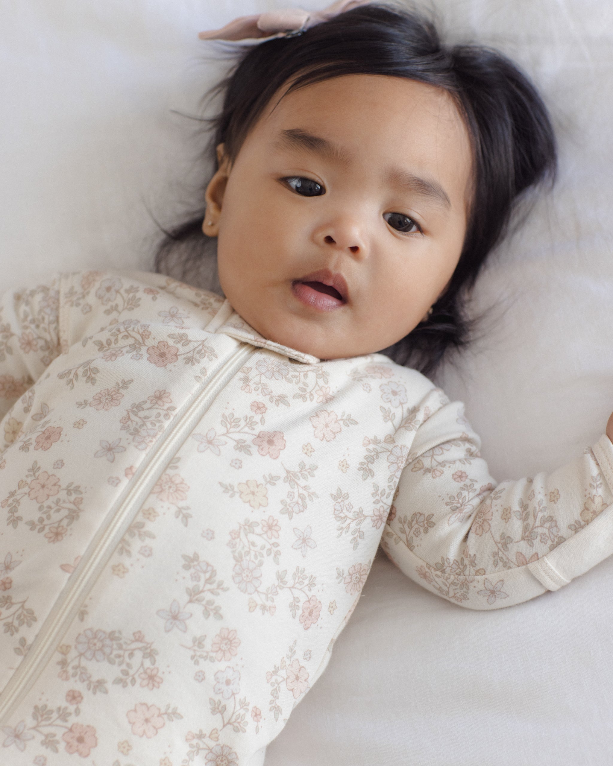 A baby lying on a white blanket, wearing a cream-colored long-sleeve sleep sack with a delicate pink and beige floral pattern.