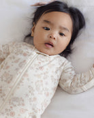 A baby lying on a white blanket, wearing a cream-colored long-sleeve sleep sack with a delicate pink and beige floral pattern.