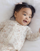 A baby lying on a soft white blanket, wearing a long-sleeve cream-colored zip-up sleeper with a delicate pink and beige floral pattern.