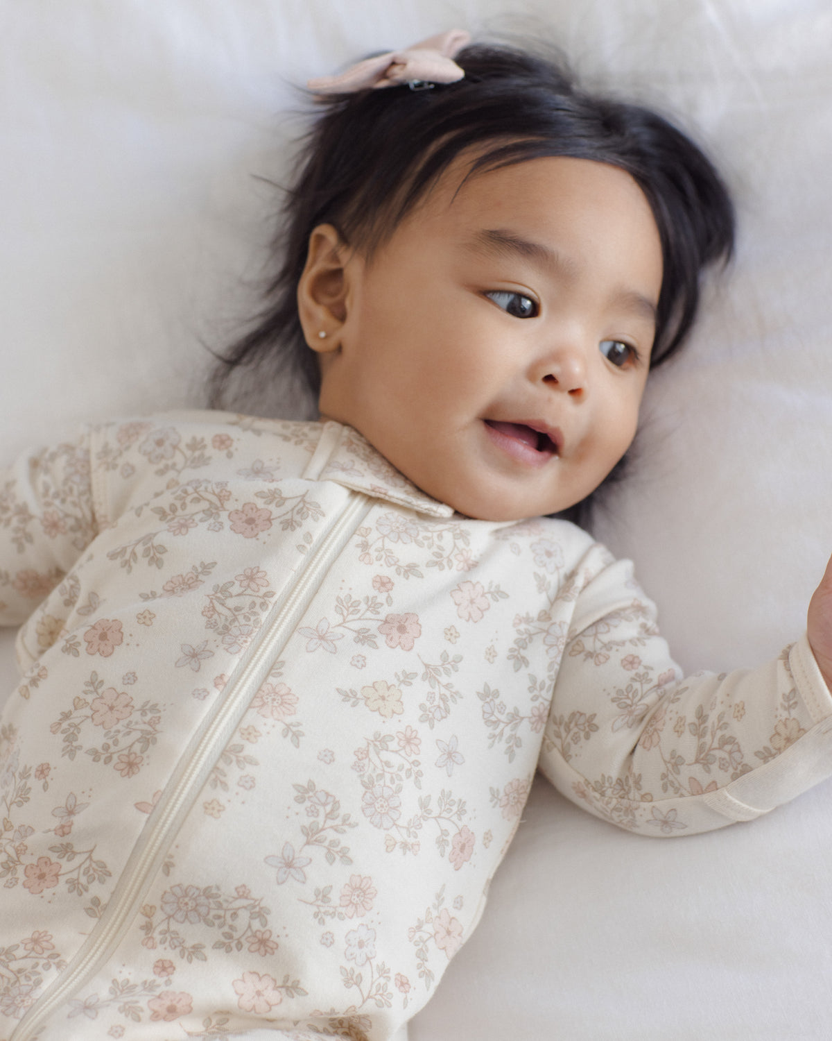 A baby lying on a soft white blanket, wearing a long-sleeve cream-colored zip-up sleeper with a delicate pink and beige floral pattern.