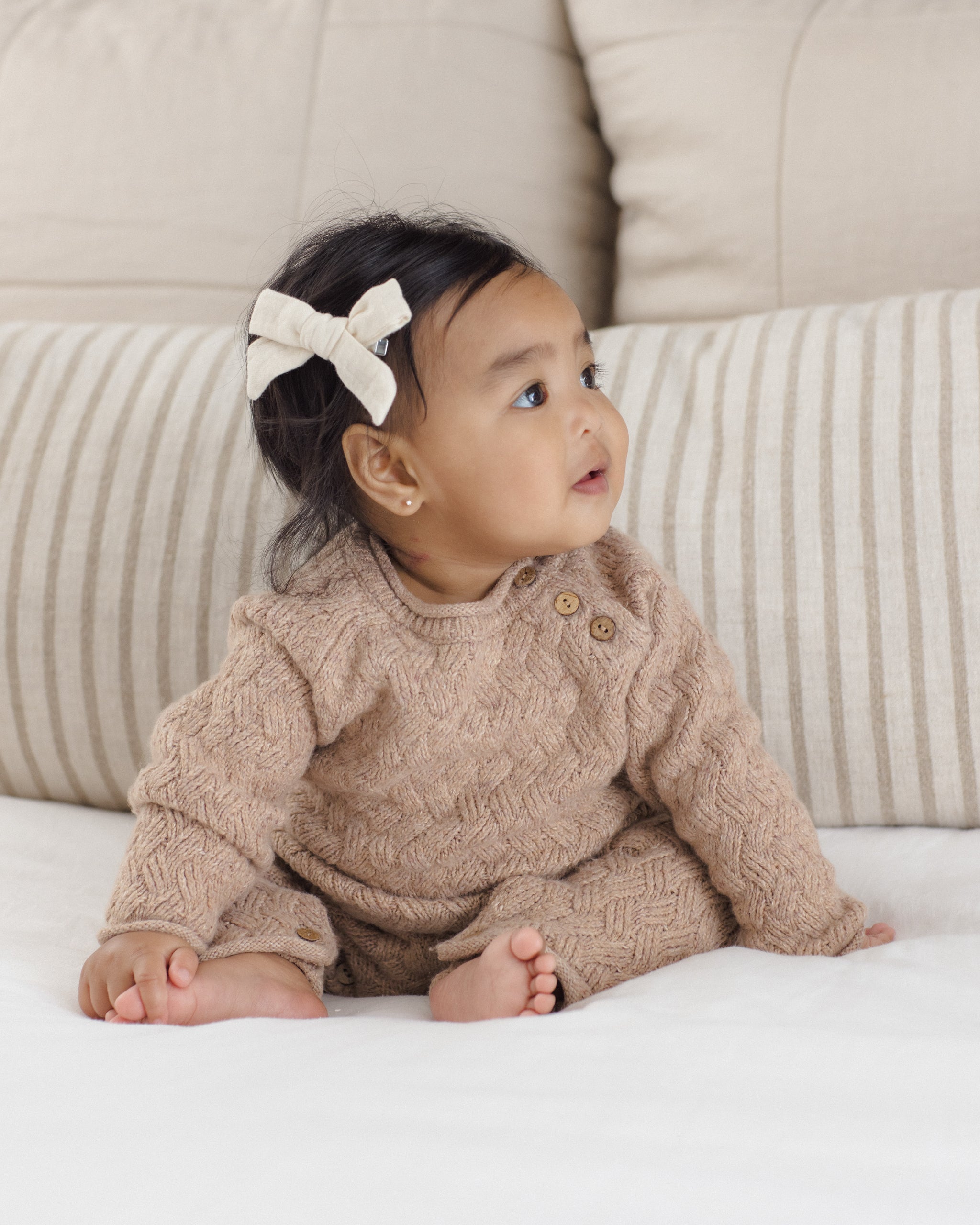 A baby wearing a beige cable-knit romper and matching hat, sitting on a white blanket in front of striped pillows.
