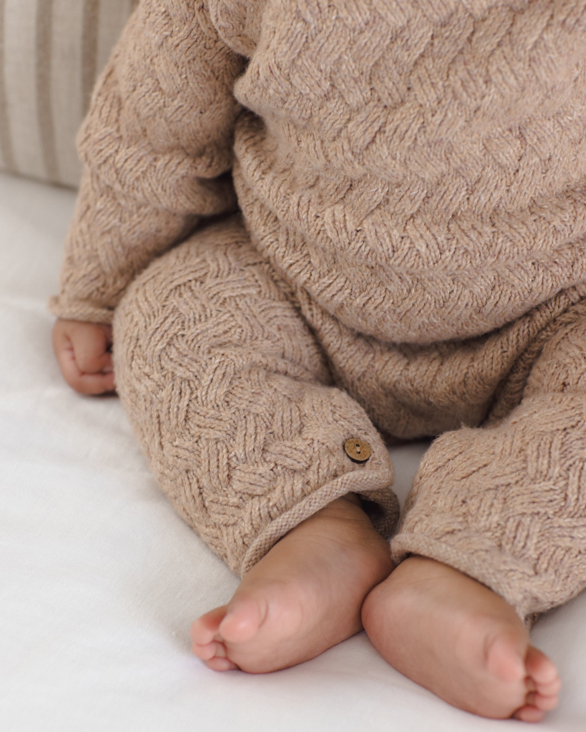 A close-up of a baby wearing a chunky cable-knit beige romper, sitting on a white blanket. The knit fabric features a detailed basketweave pattern and a small wooden button detail on the cuff of one leg.
