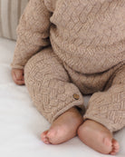 A close-up of a baby wearing a chunky cable-knit beige romper, sitting on a white blanket. The knit fabric features a detailed basketweave pattern and a small wooden button detail on the cuff of one leg.