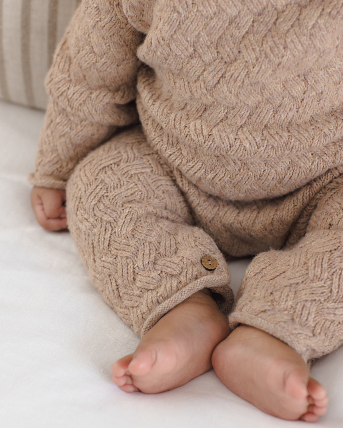 A close-up of a baby wearing a chunky cable-knit beige romper, sitting on a white blanket. The knit fabric features a detailed basketweave pattern and a small wooden button detail on the cuff of one leg.