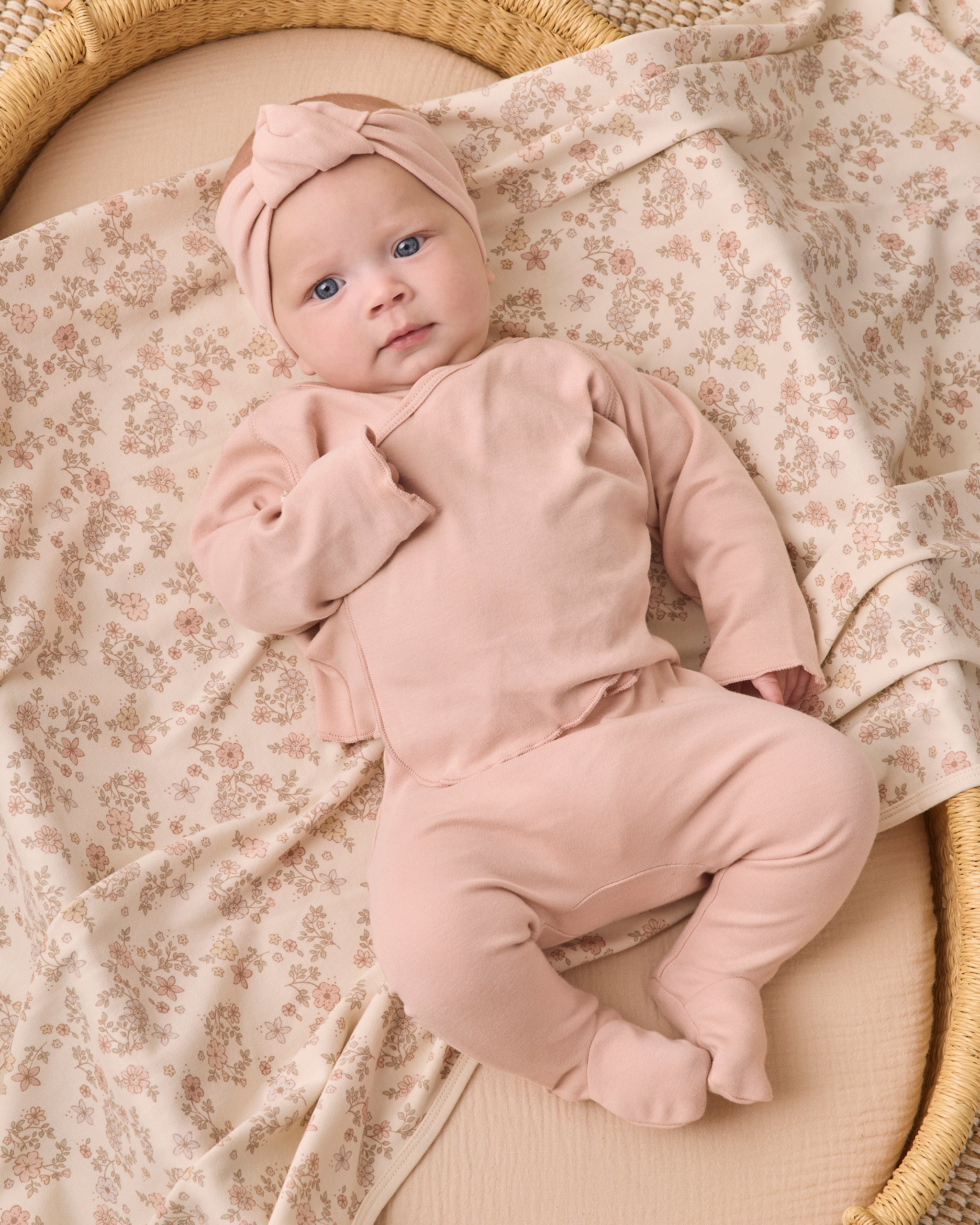 A baby lying on a woven bassinet, wearing a dusty pink footed sleeper and matching knotted headband, with a cream floral blanket underneath.