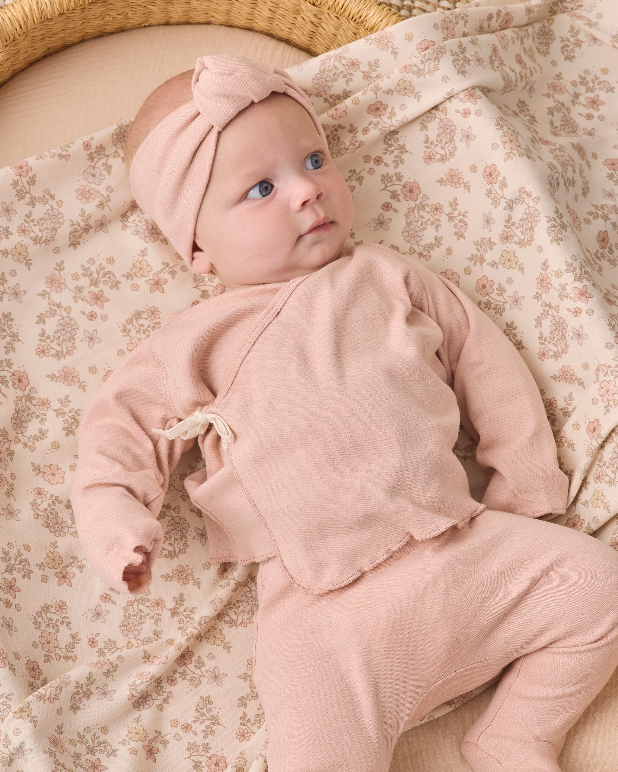 A baby lying on a floral blanket wearing a matching dusty pink two-piece outfit with a knotted headband.
