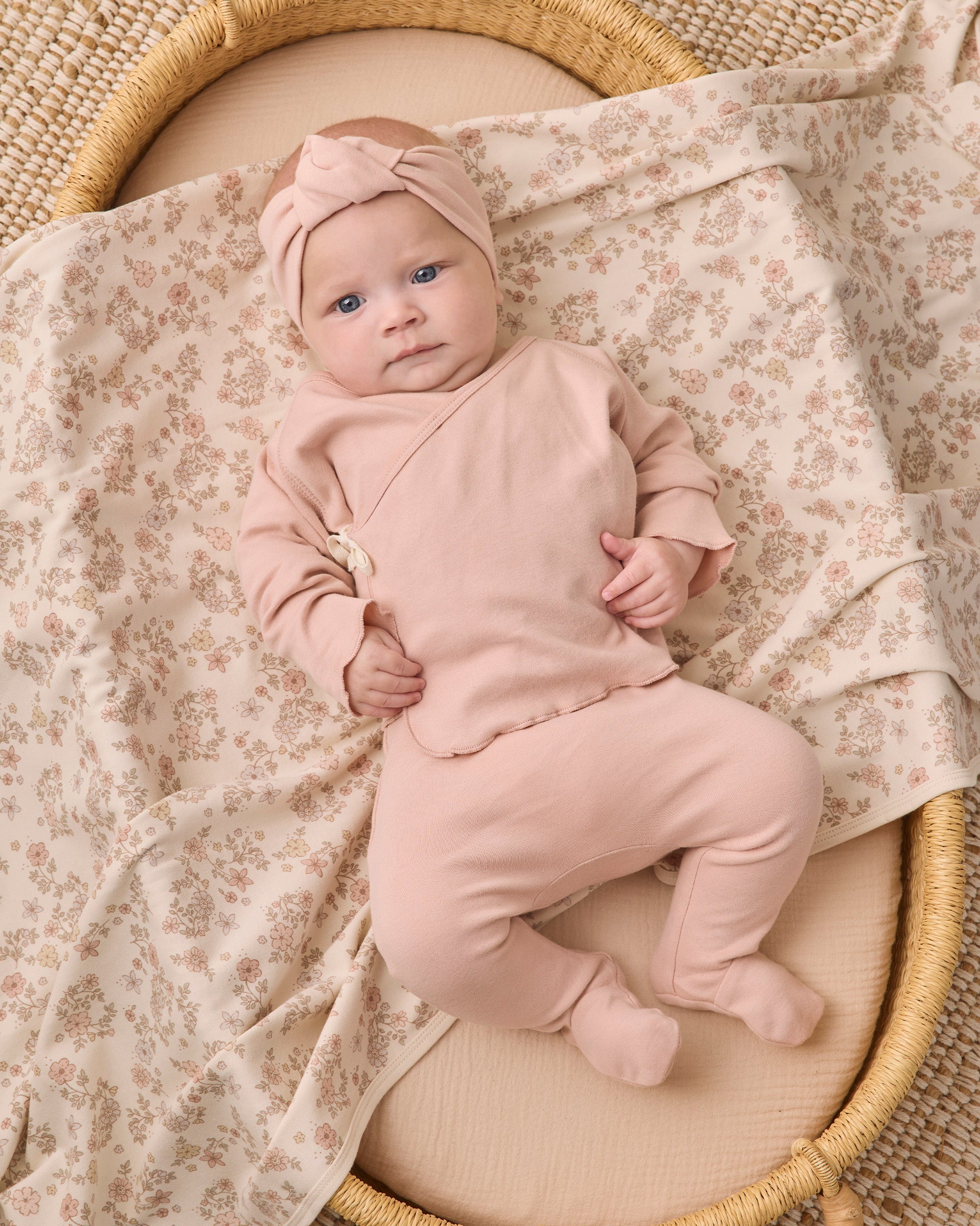 A baby wearing a dusty pink footed sleeper and matching bow headband, lying on a floral-patterned blanket inside a woven basket.