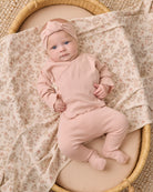 A baby wearing a dusty pink footed sleeper and matching bow headband, lying on a floral-patterned blanket inside a woven basket.