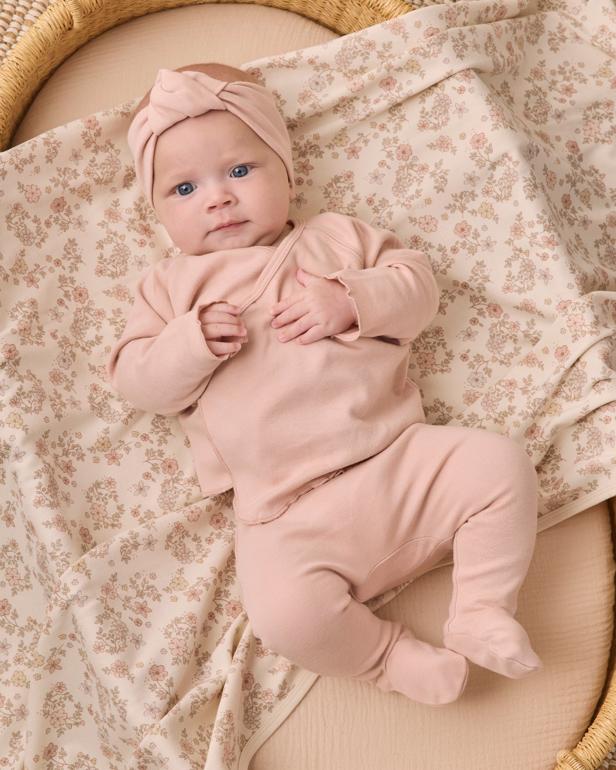 A baby lying on a floral blanket in a woven bassinet, wearing a dusty pink footed bodysuit and matching knotted headband.