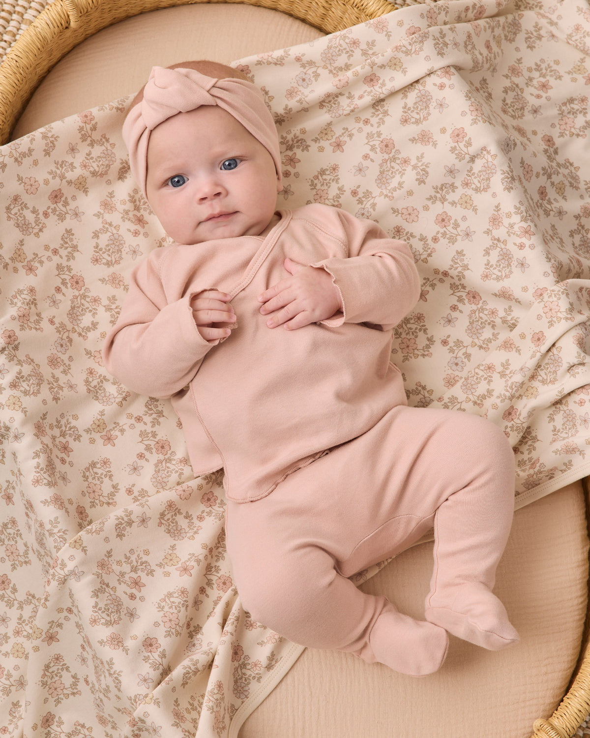 A baby lying on a floral blanket in a woven bassinet, wearing a dusty pink footed bodysuit and matching knotted headband.