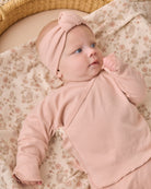 A baby wearing a dusty pink long-sleeve wrap outfit with a matching knotted headband, lying on a beige floral blanket in a woven basket.