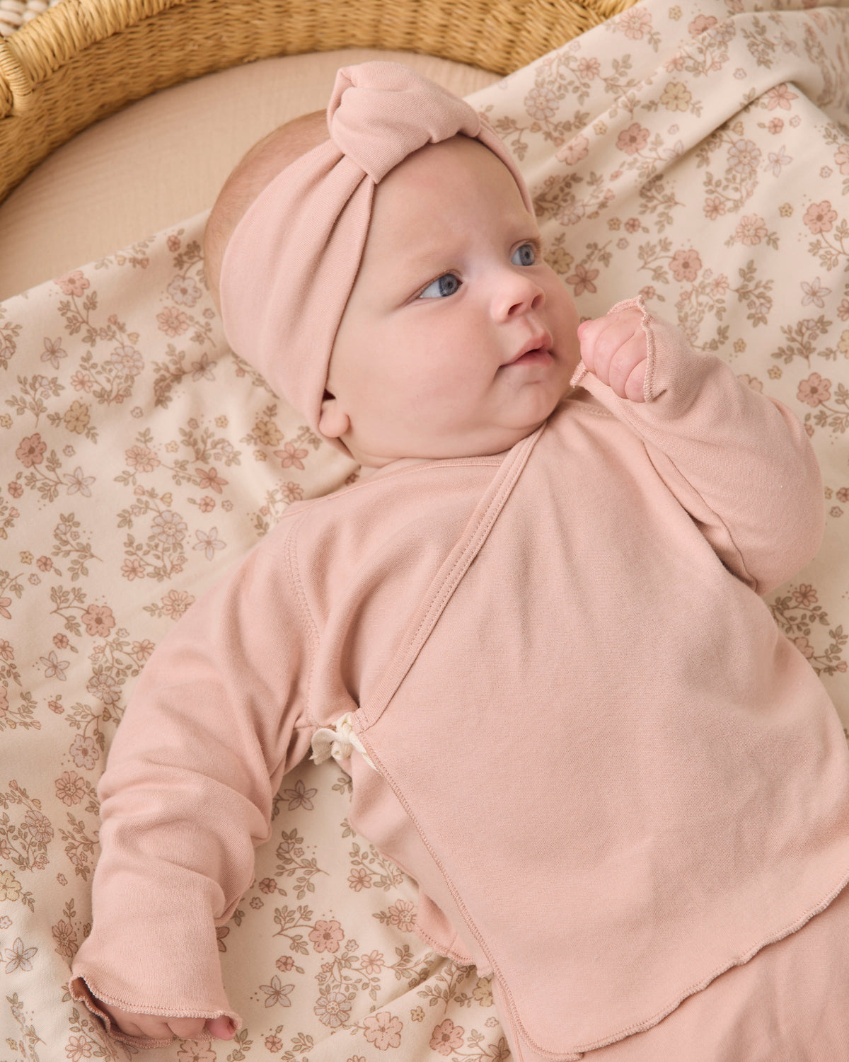 A baby wearing a dusty pink long-sleeve wrap outfit with a matching knotted headband, lying on a beige floral blanket in a woven basket.