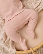 A close-up of a baby wearing dusty pink footed leggings and a matching top, lying on a cream-colored woven bassinet with floral-patterned blankets.